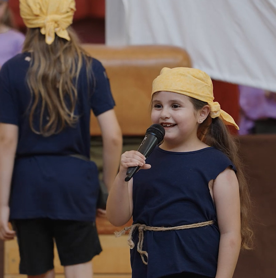 Ein junges Mädchen in einem dunkelblauen Piratenkostüm mit gelbem Kopftuch hält ein Mikrofon und spricht fröhlich auf einer Bühne. Sie trägt ein einfaches, selbstgemachtes Piratenoutfit mit einem Seil als Gürtel. Ihre langen, dunklen Haare sind zu einem Zopf gebunden. Im Hintergrund stehen weitere Kinder in Piratenkostümen vor einem großen weißen Segel als Bühnenkulisse.