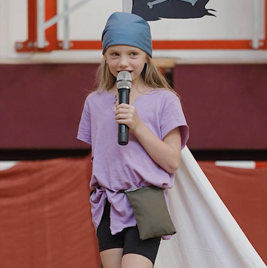Ein junges Mädchen in einem lila Piratenkostüm mit einem blauen Kopftuch steht auf einer Bühne und hält ein Mikrofon. Sie trägt ein weit geschnittenes Shirt mit kleinen Löchern, das an der Seite geknotet ist, dazu schwarze Shorts und eine kleine, olivgrüne Gürteltasche. Im Hintergrund hängt eine schwarze Piratenflagge mit einem Totenkopf und gekreuzten Knochen. Das Mädchen spricht mit einem selbstbewussten Blick ins Mikrofon und scheint eine Rolle im Stück zu spielen.
